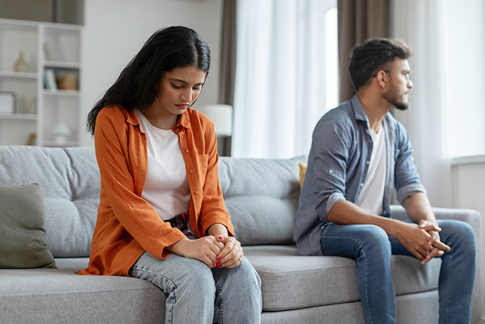 Couple sitting apart on couch, tense after truth about age deception is revealed in relationship discussion.