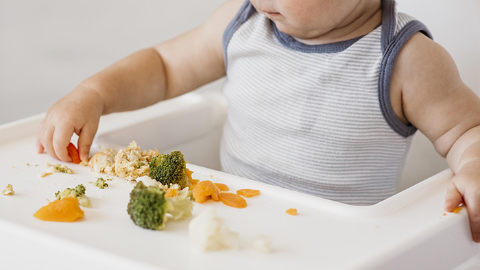 Five-month-old baby sitting in a high chair with scattered baby food, highlighting risks of pseudoscience parenting choices.