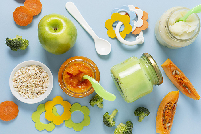 Various baby foods with fruits, vegetables, and spoons arranged on a blue surface representing infant nutrition and feeding.