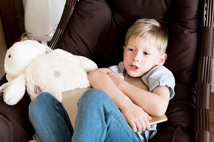 Young boy sitting in chair with tablet and stuffed animal, illustrating selfish and unloving child behavior.