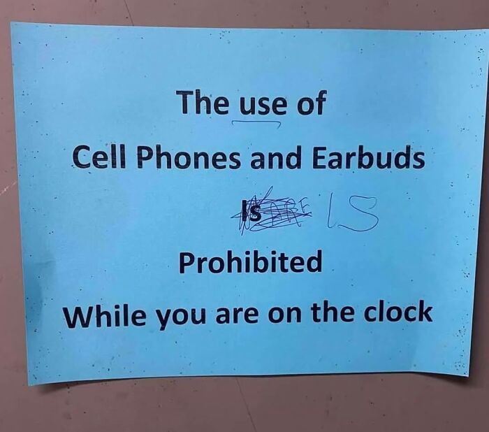 Sign on blue paper stating the use of cell phones and earbuds is prohibited while on the clock, with a handwritten correction.