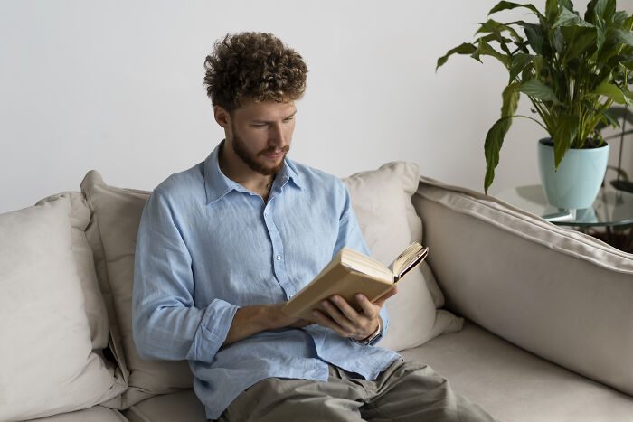 Man in a blue shirt sitting on a couch reading a book, reflecting on terrifying partner secrets discovered.