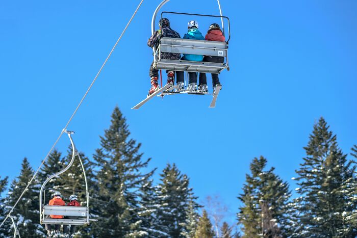 People on a ski lift surrounded by snowy pine trees under a clear blue sky, evoking a scary mountain experience.