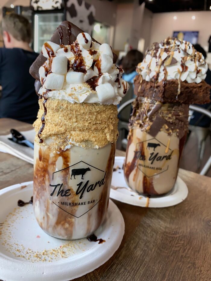 Two decadent milkshakes topped with whipped cream, marshmallows, brownies, and chocolate drizzle, perfect dishes for foodies.