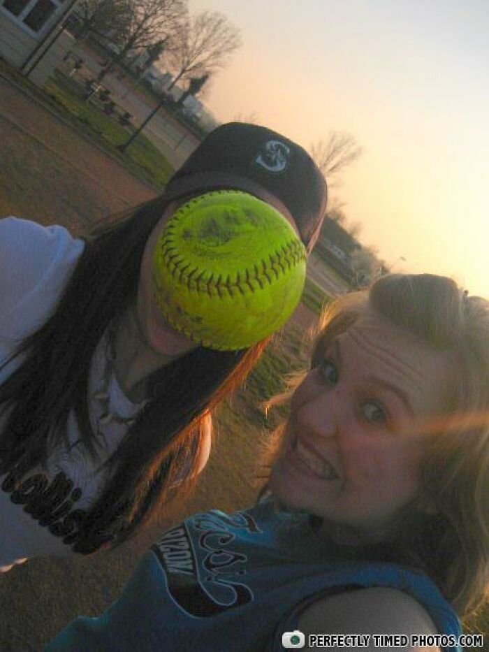 Two girls outdoors with a softball perfectly timed to cover one girl’s face in a perfectly timed photo.