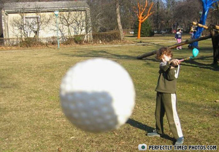 Blurred golf ball in the foreground with a child swinging a club in the background, a perfectly timed photo outdoors.