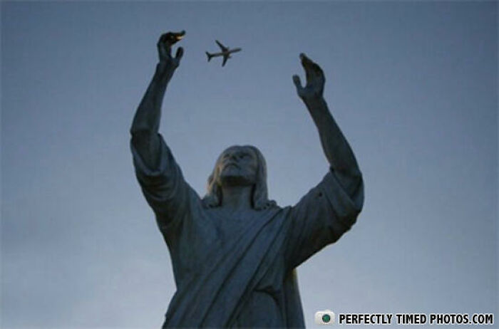 Statue with arms raised appearing to catch an airplane flying overhead in a perfectly timed photo.
