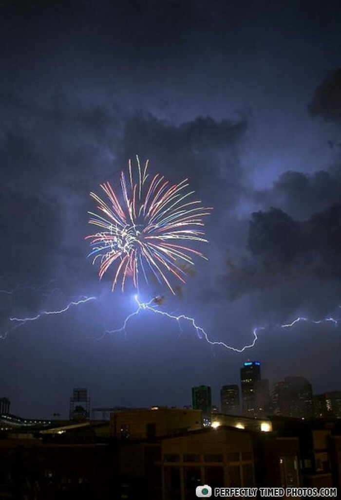 Fireworks burst in the sky perfectly timed with a lightning strike over a cityscape at night in an impressive photo.