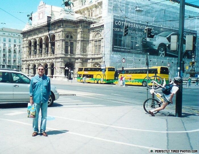 Man standing still on busy city street while cyclist is captured falling off bike in a perfectly timed photo.