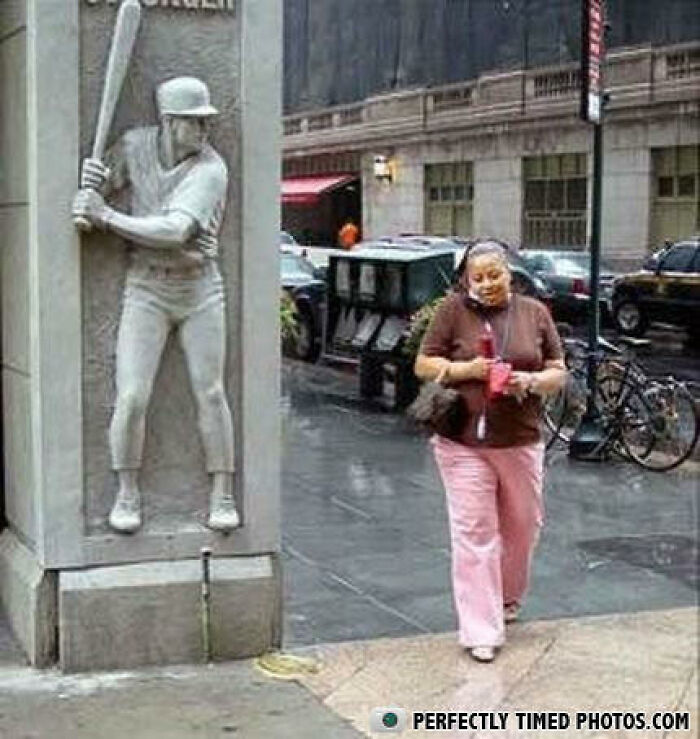 Woman walking on city street perfectly timed with baseball statue preparing to swing bat on corner.