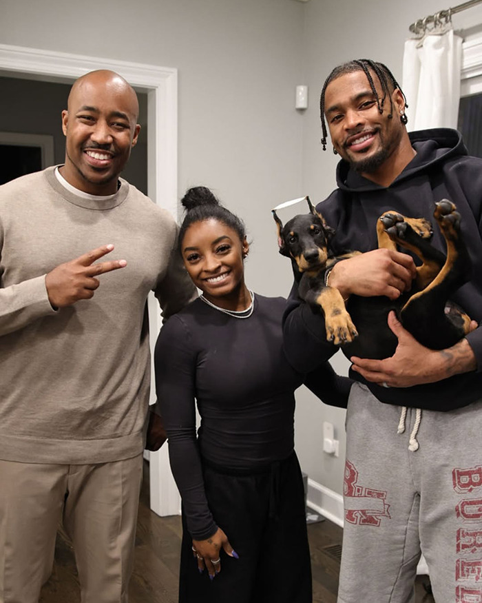Simone Biles standing between two men indoors, while one man holds a dog with cropped ears in his arms.