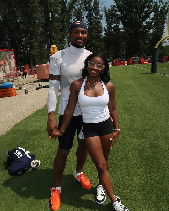 Simone Biles smiling outdoors on a sports field, wearing athletic attire and standing with a man in sporty clothes.