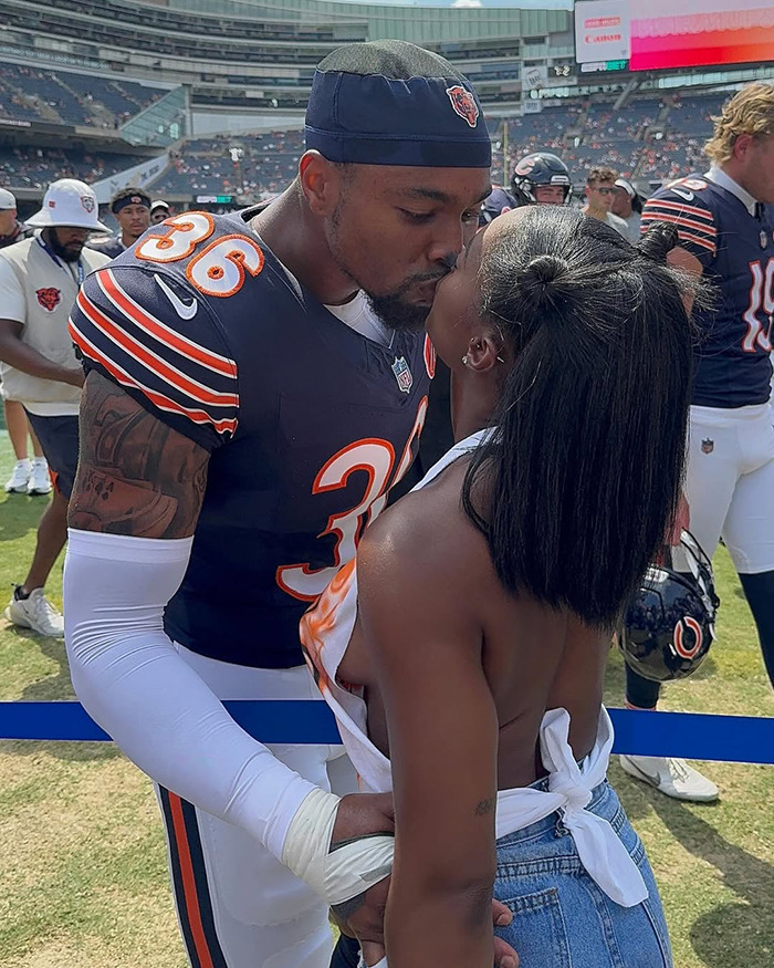 Chicago Bears player in uniform kissing a woman on the field after a game, with teammates in the background.
