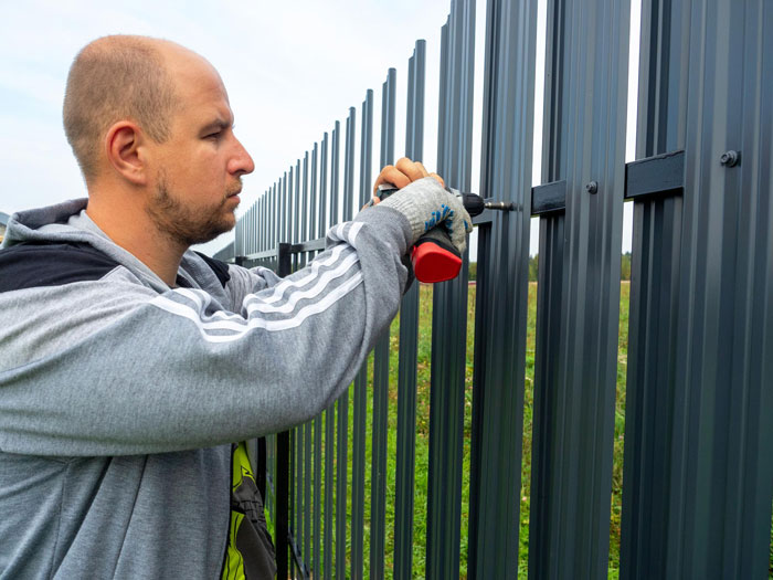 Man wearing gloves using a drill to install a metal fence outdoors, illustrating Karen Petty fence revenge concept.
