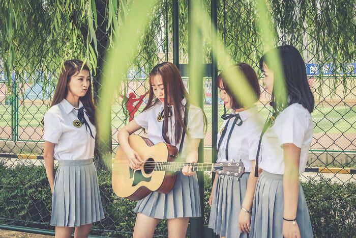 Four young women in school uniforms, one playing guitar, outdoors near a fence, capturing unresolved mysteries atmosphere.
