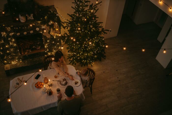 Family enjoying a cozy Christmas dinner beside a decorated tree with festive string lights and holiday treats.