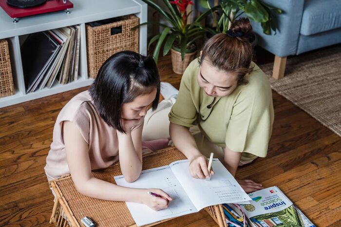 Two women sitting on floor, engaging in modern parenting trends by helping with homework together at home.
