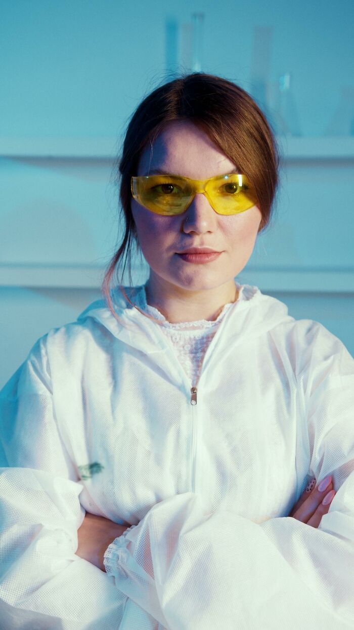 Woman wearing yellow protective glasses and a white suit, confidently standing with arms crossed for sneaky acts of revenge.