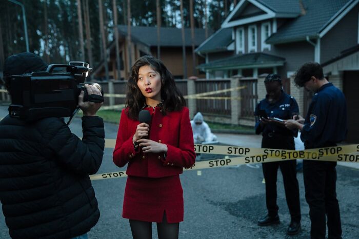 Reporter in red suit covering disturbing truths at a crime scene with police and caution tape in the background.