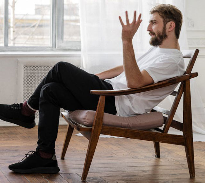 Young man sitting in a wooden chair indoors, gesturing with his hand, reflecting on family disownment and shame.