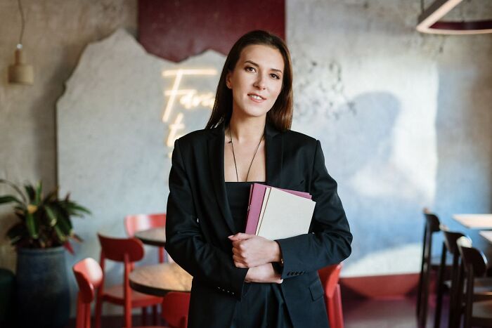 Young professional woman in black blazer holding folders, standing confidently in a modern cafe setting with clever and sassy vibes.