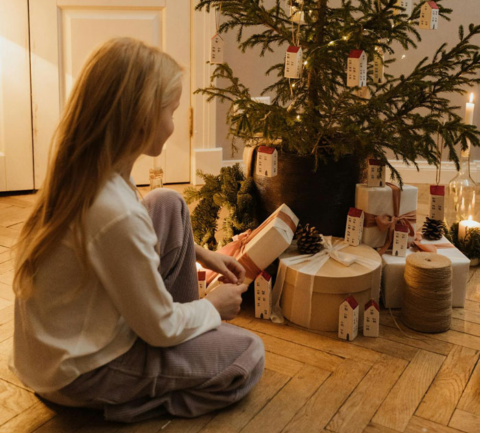 Young girl sitting by Christmas tree holding a gift in a cozy room with wrapped presents and festive decorations. Young girl sitting by Christmas tree holding a gift in a cozy room with wrapped presents and festive decorations.