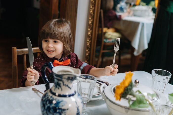 Young child at dining table holding knife and fork, displaying pure entitlement with an excited expression.