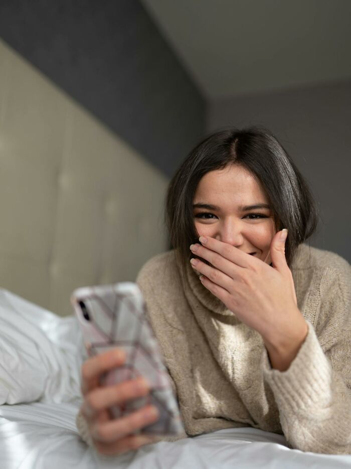 Young woman in a cozy sweater laughing sneakily while holding her phone, hinting at brilliantly sneaky acts of revenge.