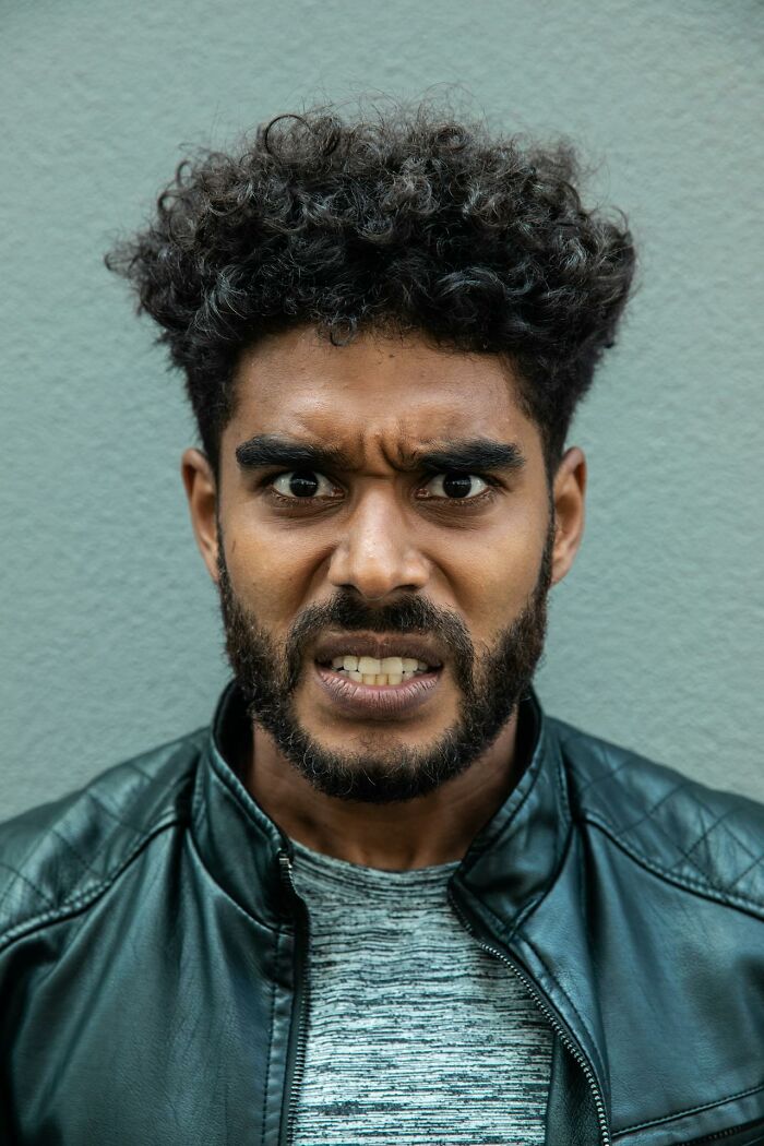 Man with curly hair and beard showing anger and entitlement, wearing a leather jacket against a gray wall background