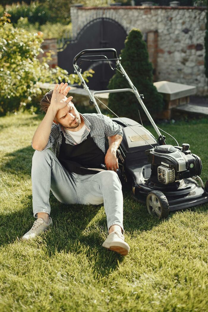 Man sitting on grass next to lawn mower, shielding eyes from sun, illustrating brilliantly sneaky acts of revenge concept.