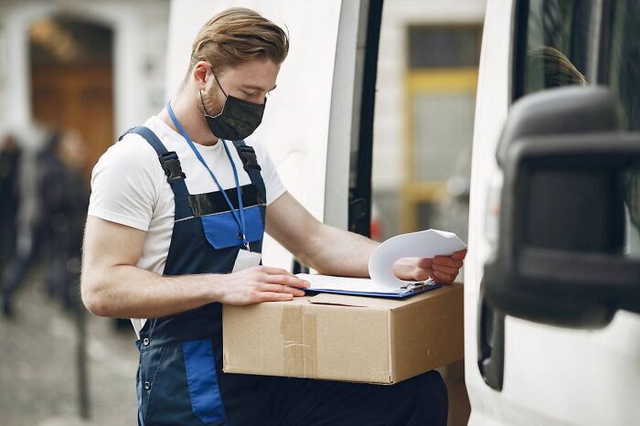 Postal worker in uniform wearing a mask, checking a package and paperwork while standing beside a delivery van.