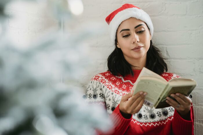 Woman wearing a Santa hat and festive sweater reading a book, highlighting the concept of horrible Christmas gifts.