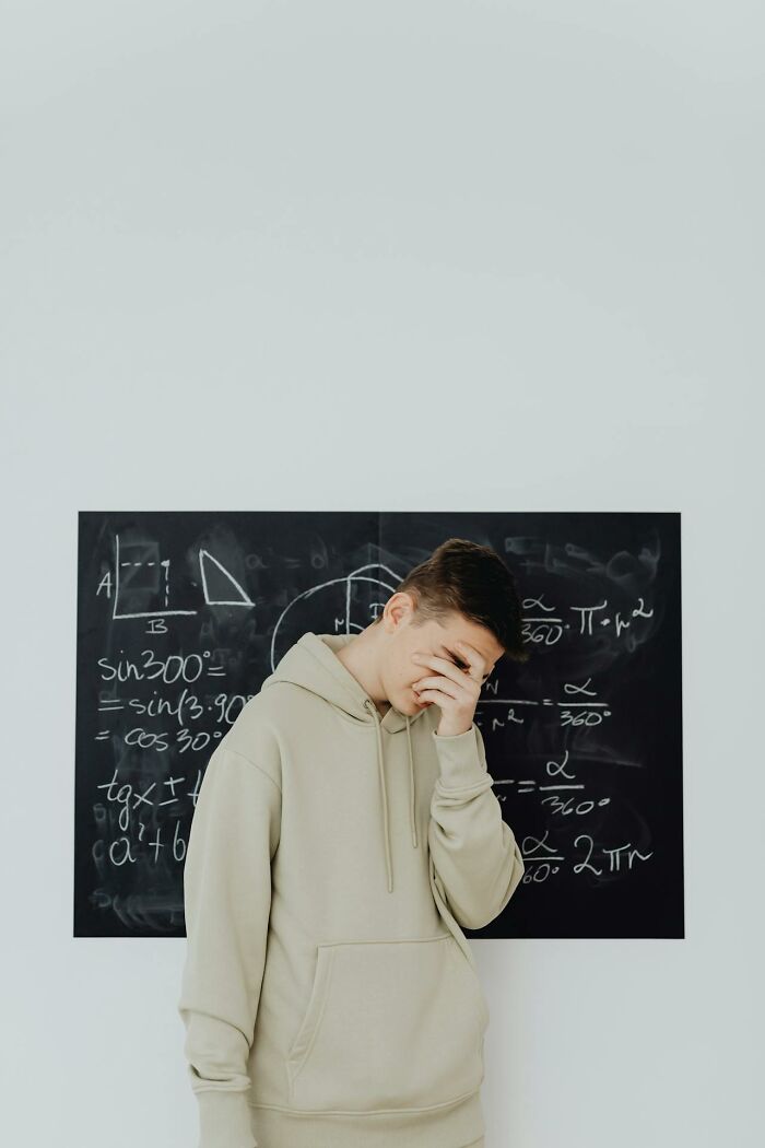 Young man in beige hoodie covering face in front of a blackboard full of math equations, showing a glitch in the system moment.