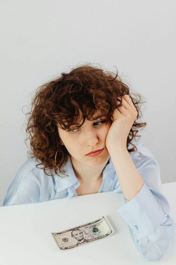 Young woman with curly hair looking distressed and tired, resting her head on her hand near a five-dollar bill.
