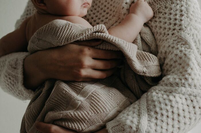 Adult wearing a knitted cream sweater holding a baby wrapped in a beige knitted blanket, evoking comforting yet unsettling feelings.