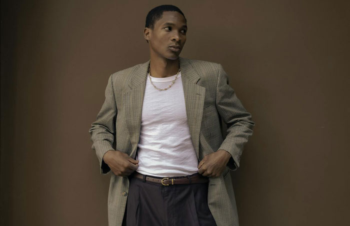 Young Black man transforming his look wearing a checked blazer, white shirt, and layered gold chain against a brown backdrop.