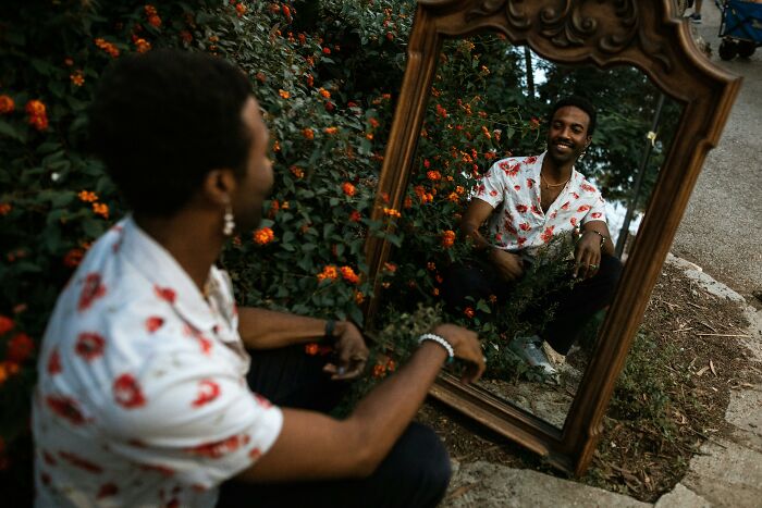 Man wearing floral shirt sits outdoors looking at himself in an ornate mirror surrounded by flowers and greenery, reflecting holiday gifts.