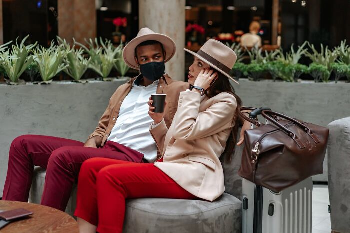 Two travelers wearing hats and masks sitting in an airport lounge with luggage and a coffee cup nearby