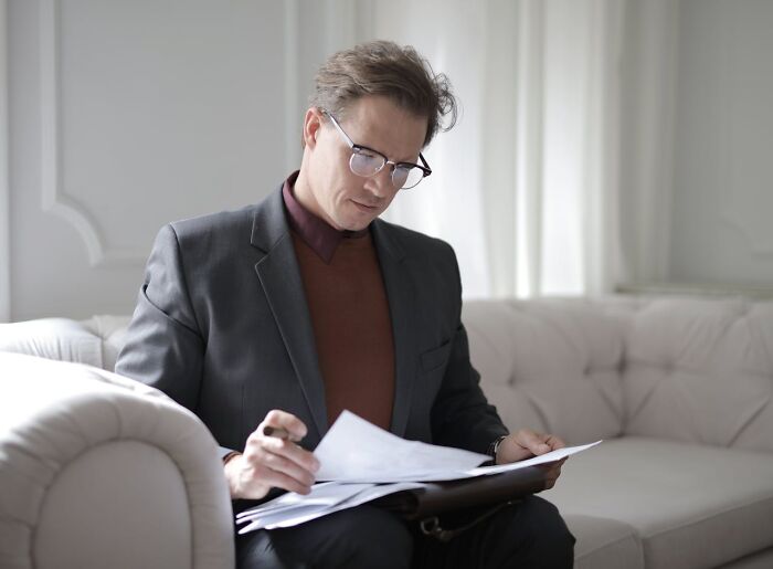 Man in glasses and suit sitting on a couch reviewing disturbing truths on papers in a bright living room setting.