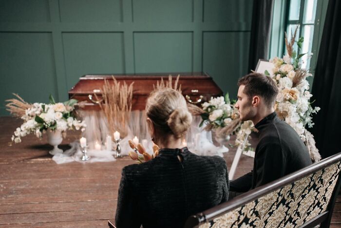 Two people sitting in a funeral home with a coffin and floral arrangements during an unexpected funeral moment.