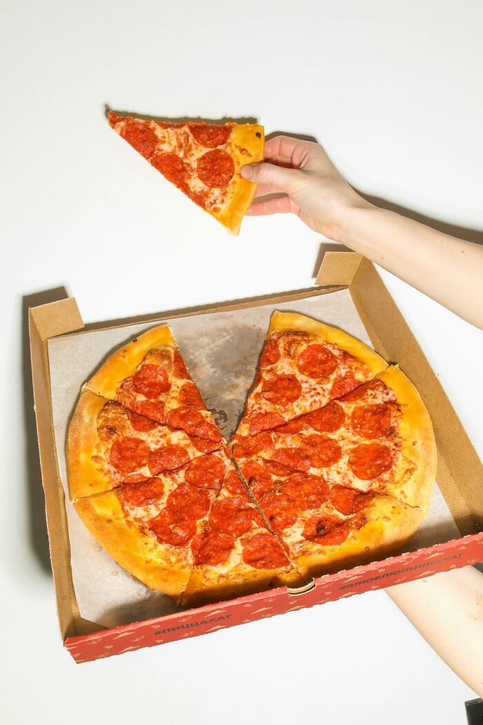 Hand holding a slice of pepperoni pizza above a pizza box showing a glitch in the system with uneven pizza slices.