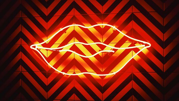 Neon lips sign glowing bright orange against a geometric patterned wall, illustrating facial recognition challenge. Neon lips sign glowing bright orange against a geometric patterned wall, illustrating facial recognition challenge.