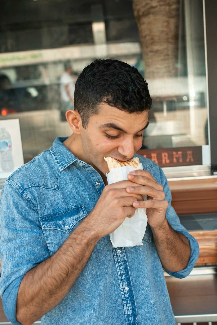 Man in denim shirt eating a sandwich outside, capturing a casual moment related to crazy airport stories and travel experiences.