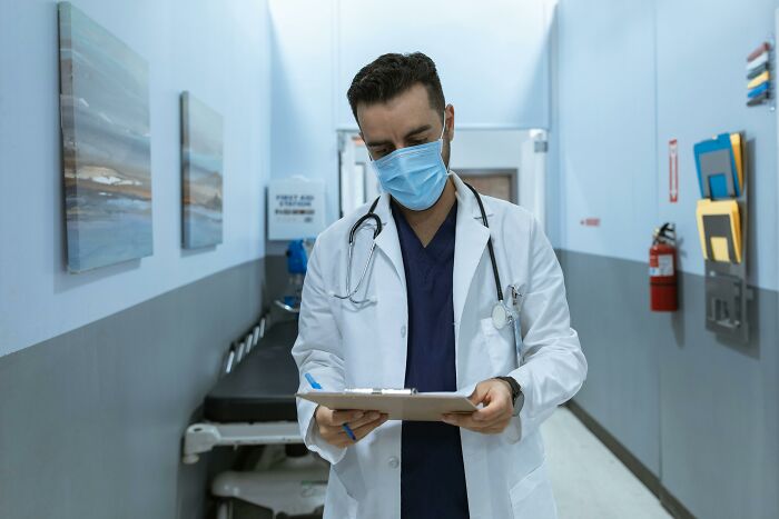 Male doctor wearing mask and white coat in hospital hallway, reviewing patient chart related to men missed delivery babies.