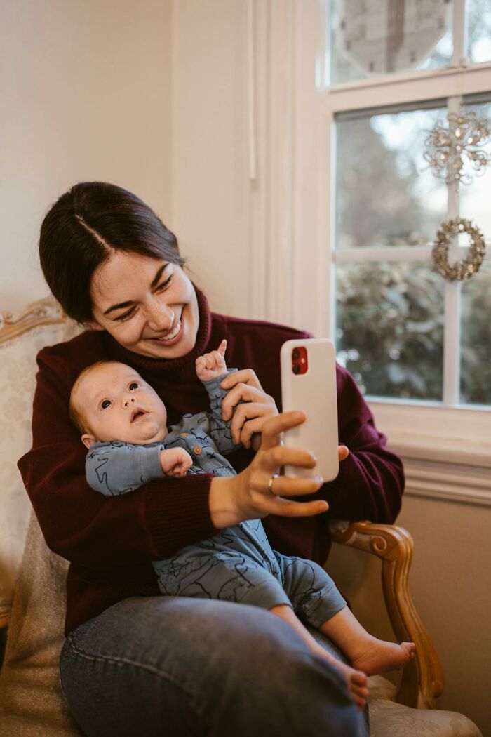 Mother holding baby while taking a selfie, illustrating modern parenting trends in a cozy home setting by the window.