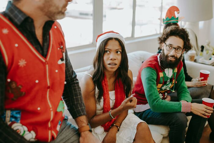 Three people wearing festive holiday outfits and hats, reacting to horrible Christmas gifts during a gathering.