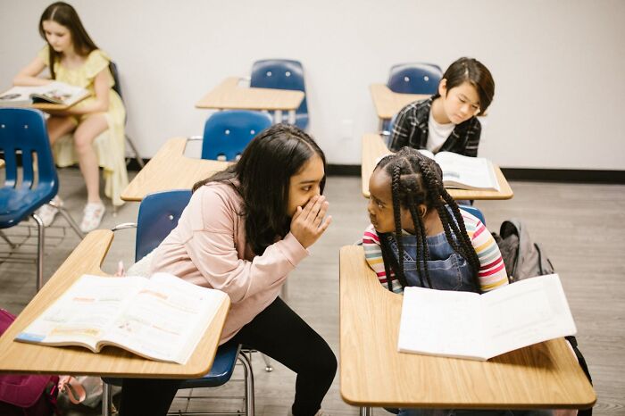 Two schoolgirls whispering across desks with open books, capturing funny, clever and sassy moments in the heat of the moment.