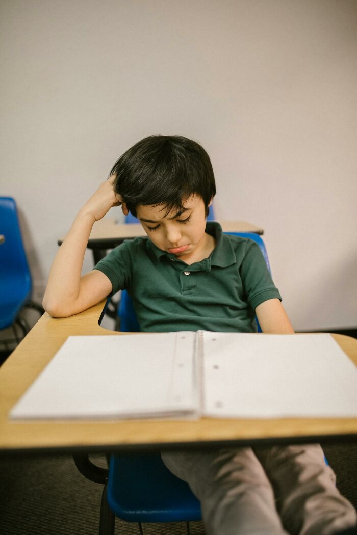 Young boy in classroom looking frustrated while sitting at desk with open notebook, showing sneaky acts of revenge concept.