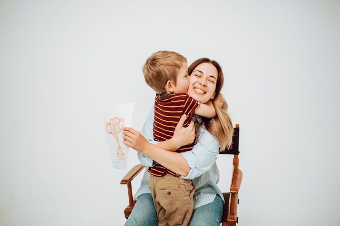 Mother sitting on a chair hugging her young son who is holding a drawing, representing modern parenting trends.