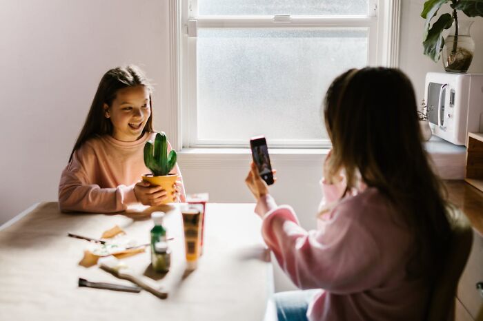 Two girls sitting at a table with craft supplies, one holding a cactus plant while the other takes a photo, illustrating modern parenting trends.